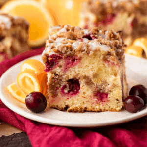 A slice of crumb-topped coffee cake with visible cranberries and drizzle of icing, served on a plate with fresh orange slices and whole cranberries, set on a red cloth.