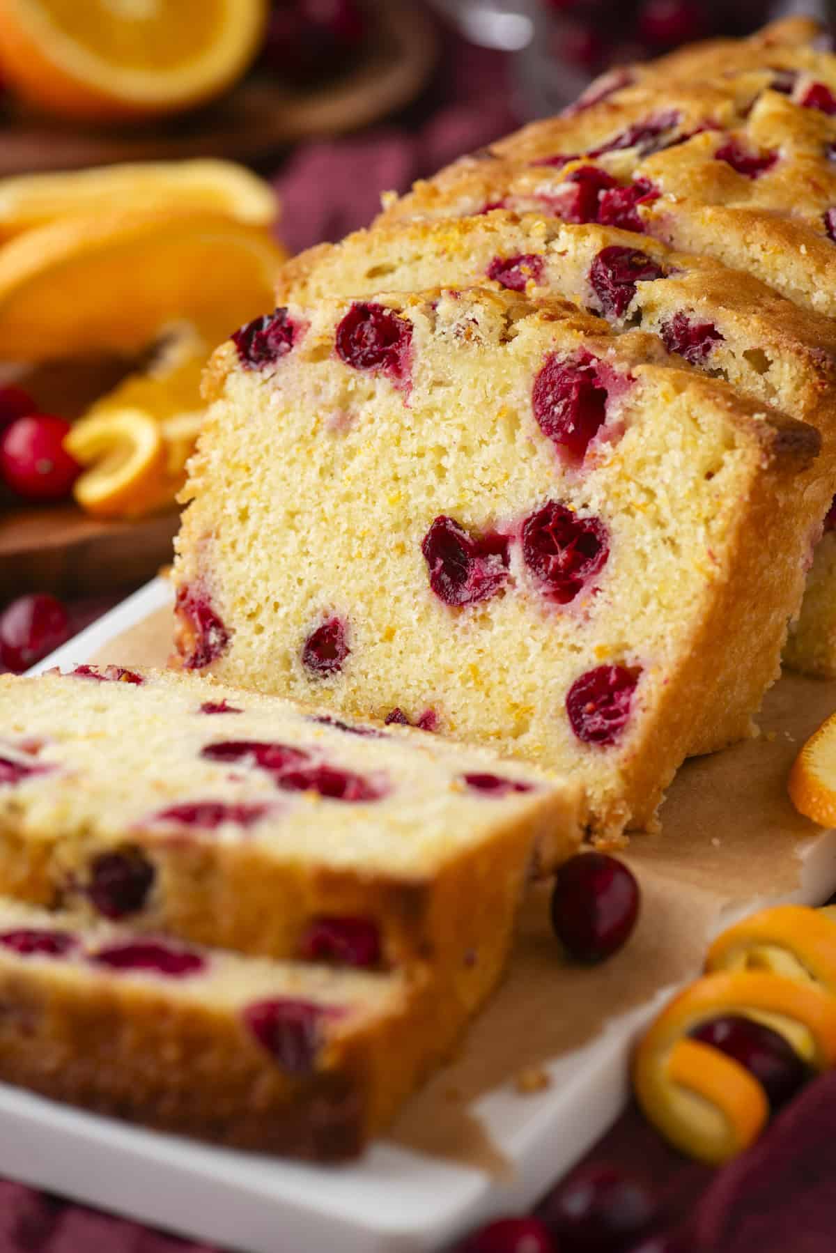 A sliced loaf of cranberry orange bread sits on a white platter, showing a golden crumb with visible cranberries. Fresh cranberries and orange slices are scattered around the bread.