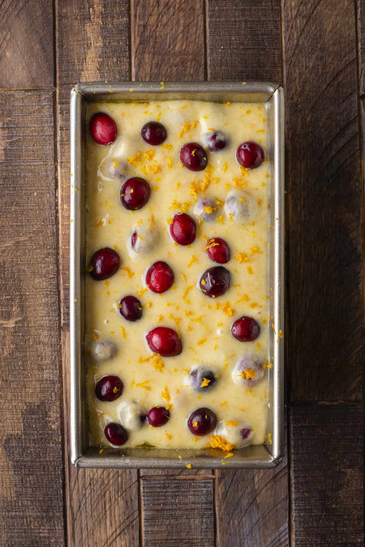 A metal loaf pan filled with unbaked batter, dotted with whole cranberries and orange zest, sits on a wooden surface.