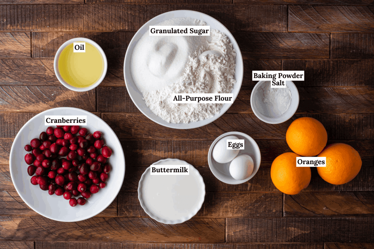 Top-down view of labeled baking ingredients: oil, cranberries, buttermilk, granulated sugar, all-purpose flour, baking powder, salt, eggs, and whole oranges, all arranged on a wooden surface.