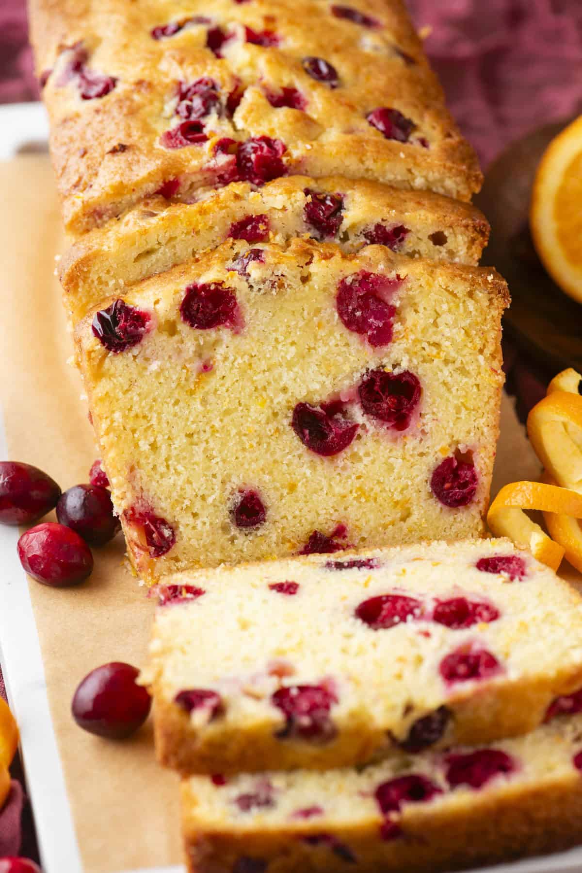 A close-up of a sliced cranberry orange loaf cake on parchment paper, with visible cranberries inside the moist yellow cake. Fresh cranberries and orange slices are nearby.