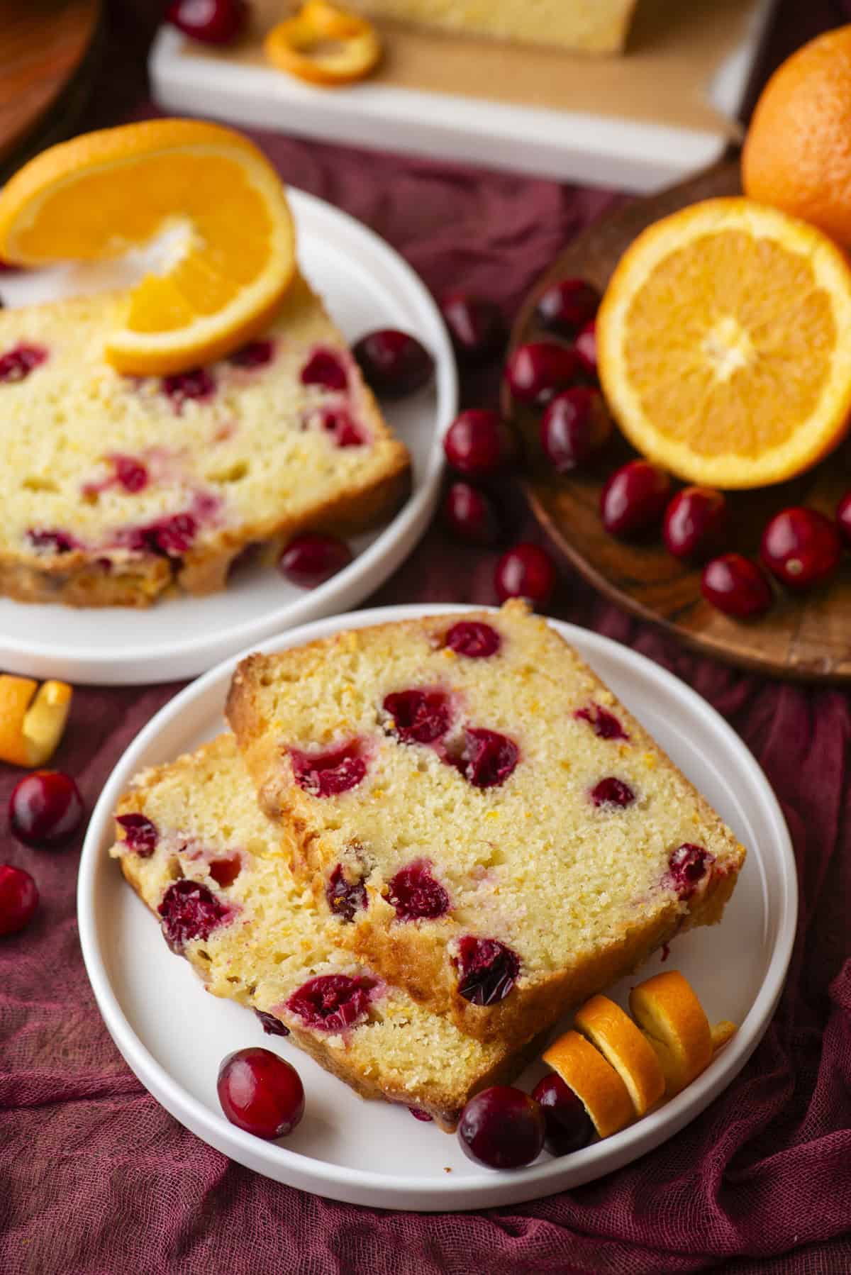 Two slices of cranberry orange bread on a white plate, garnished with fresh cranberries and orange slices, surrounded by whole oranges and cranberries on a burgundy cloth.