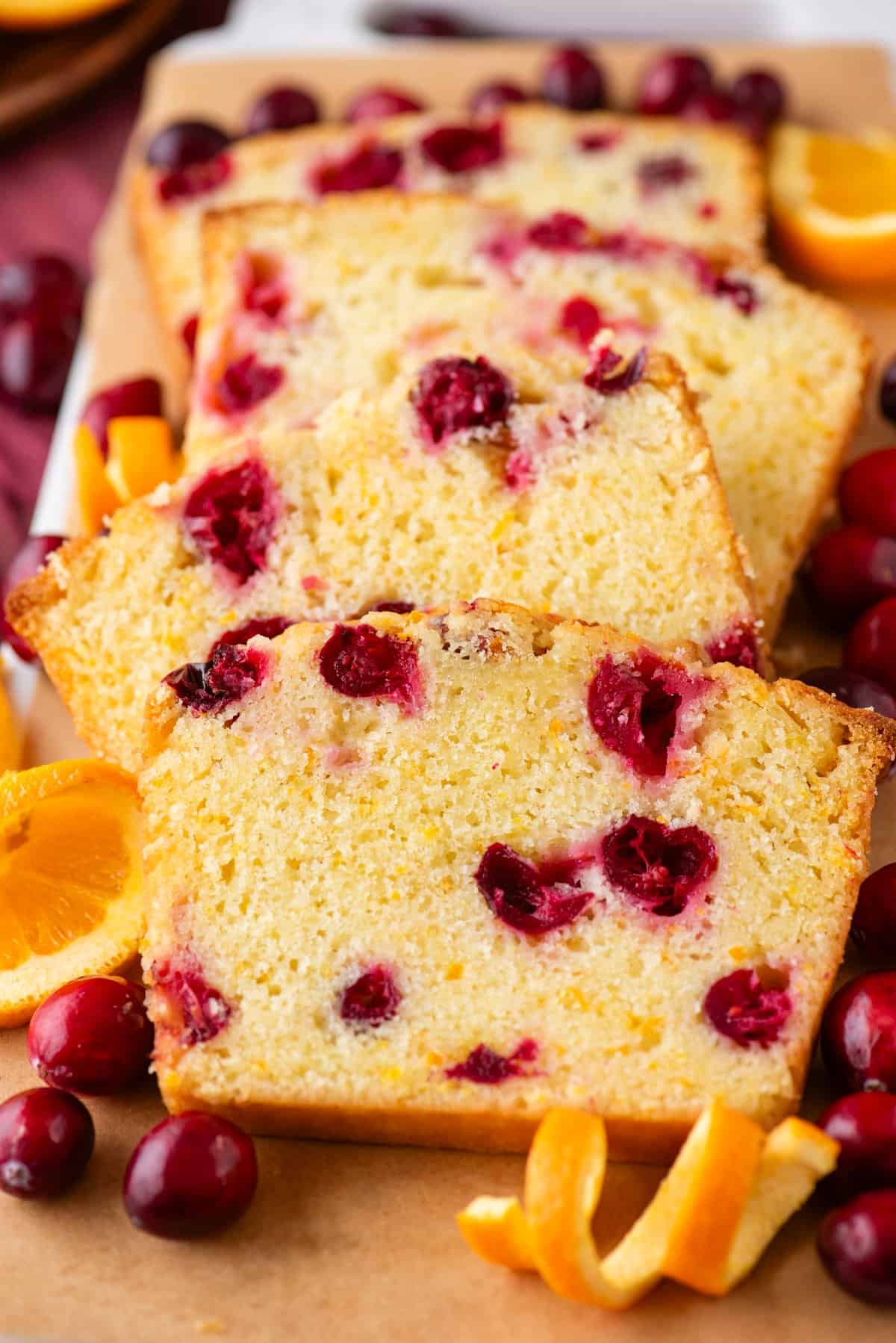 Sliced cranberry orange bread on parchment paper, garnished with whole cranberries and orange slices. The bread is golden with visible pieces of cranberry and orange zest.