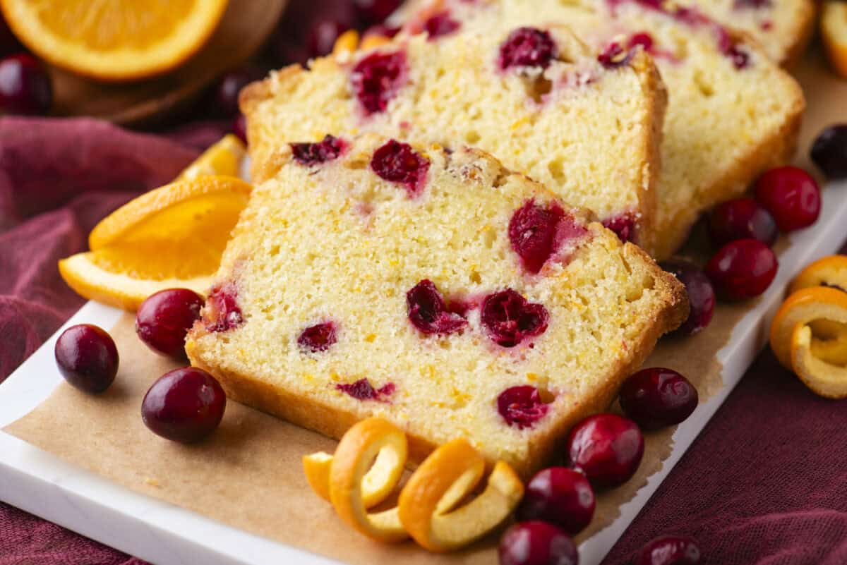 Two slices of cranberry orange bread sit on a cutting board, surrounded by fresh cranberries, orange slices, and orange zest curls, all atop a burgundy cloth. The bread is golden and dotted with cranberries.