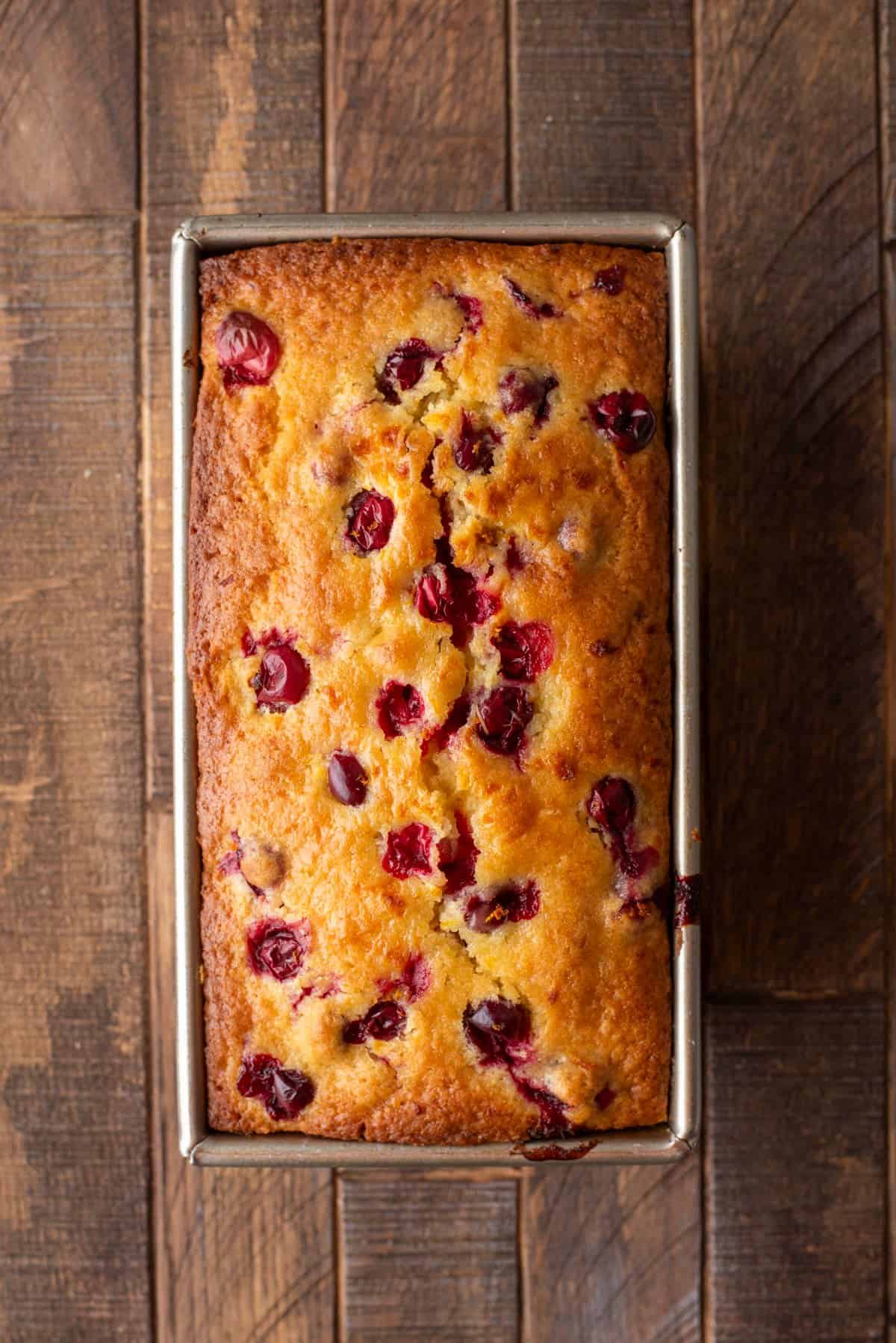A golden-brown loaf of cranberry bread in a rectangular metal pan sits on a rustic wooden surface, with bright red cranberries visible on top.