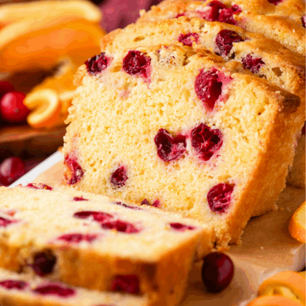 Sliced cranberry orange bread sits on a cutting board, with visible cranberries and orange pieces. Orange slices and whole cranberries are scattered nearby for decoration.