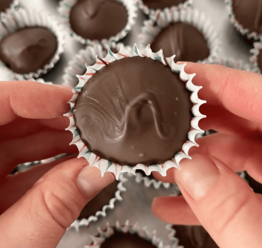 A close-up of hands holding a chocolate candy in a striped paper cup, with more chocolates in similar cups visible in the background.
