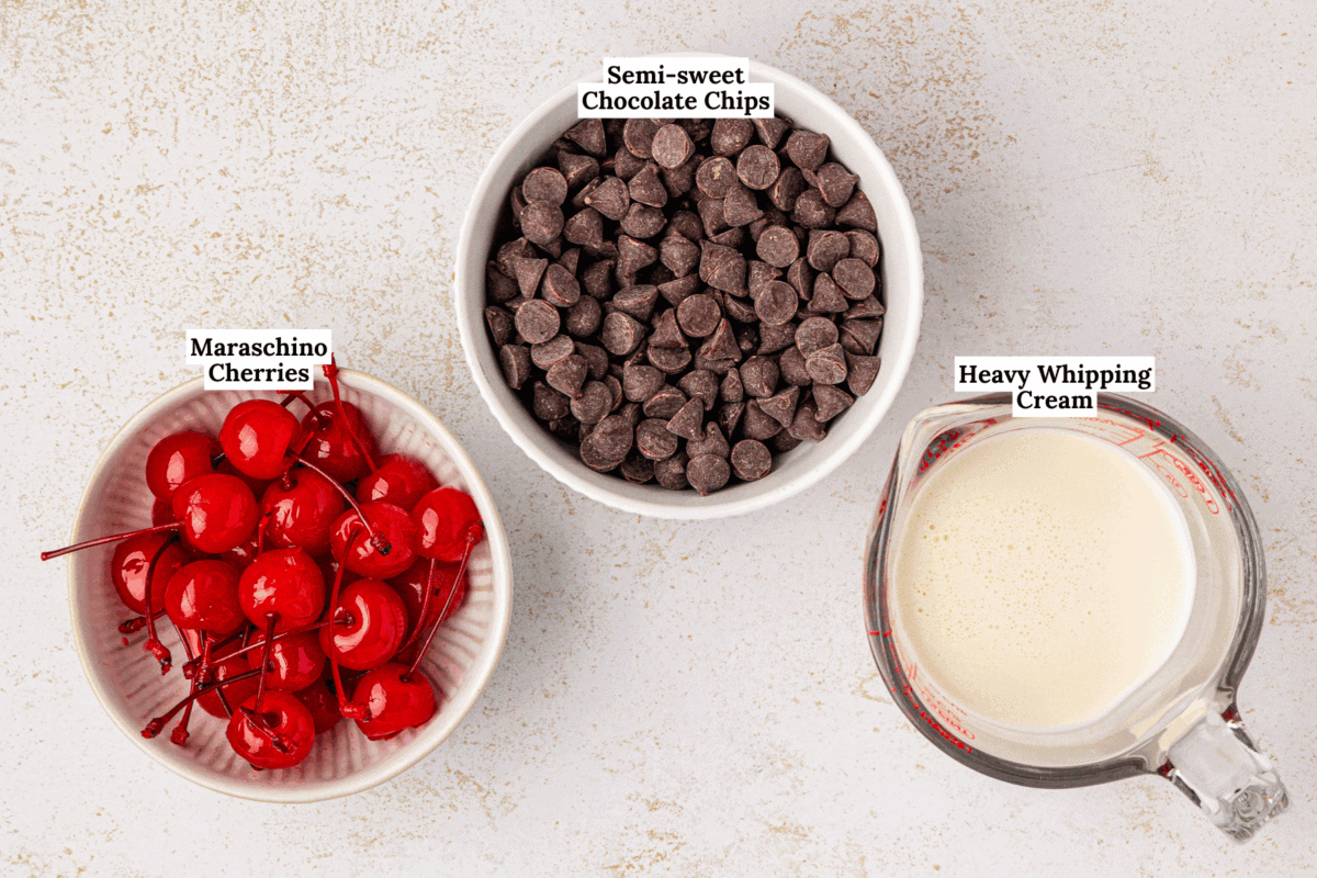 A bowl of maraschino cherries, a bowl of semi-sweet chocolate chips, and a measuring cup of heavy whipping cream are arranged on a light surface, each labeled with its name.