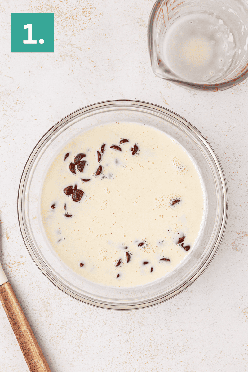 A glass bowl filled with a creamy mixture and chocolate chips sits on a light countertop. A measuring cup with some liquid and a wooden-handled spatula are nearby. A green box with the number 1 is in the top left corner.