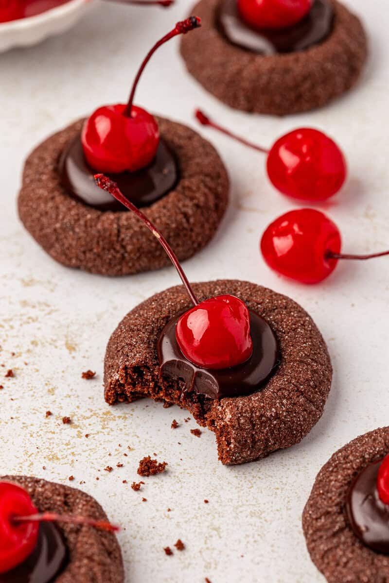 Chocolate thumbprint cookies filled with chocolate ganache and topped with maraschino cherries. One cookie has a bite taken out, showing the soft texture. Other whole cookies and loose cherries are nearby on a white surface.