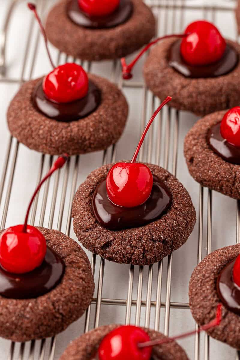 Chocolate thumbprint cookies topped with glossy chocolate ganache and bright red maraschino cherries, arranged neatly on a metal cooling rack.