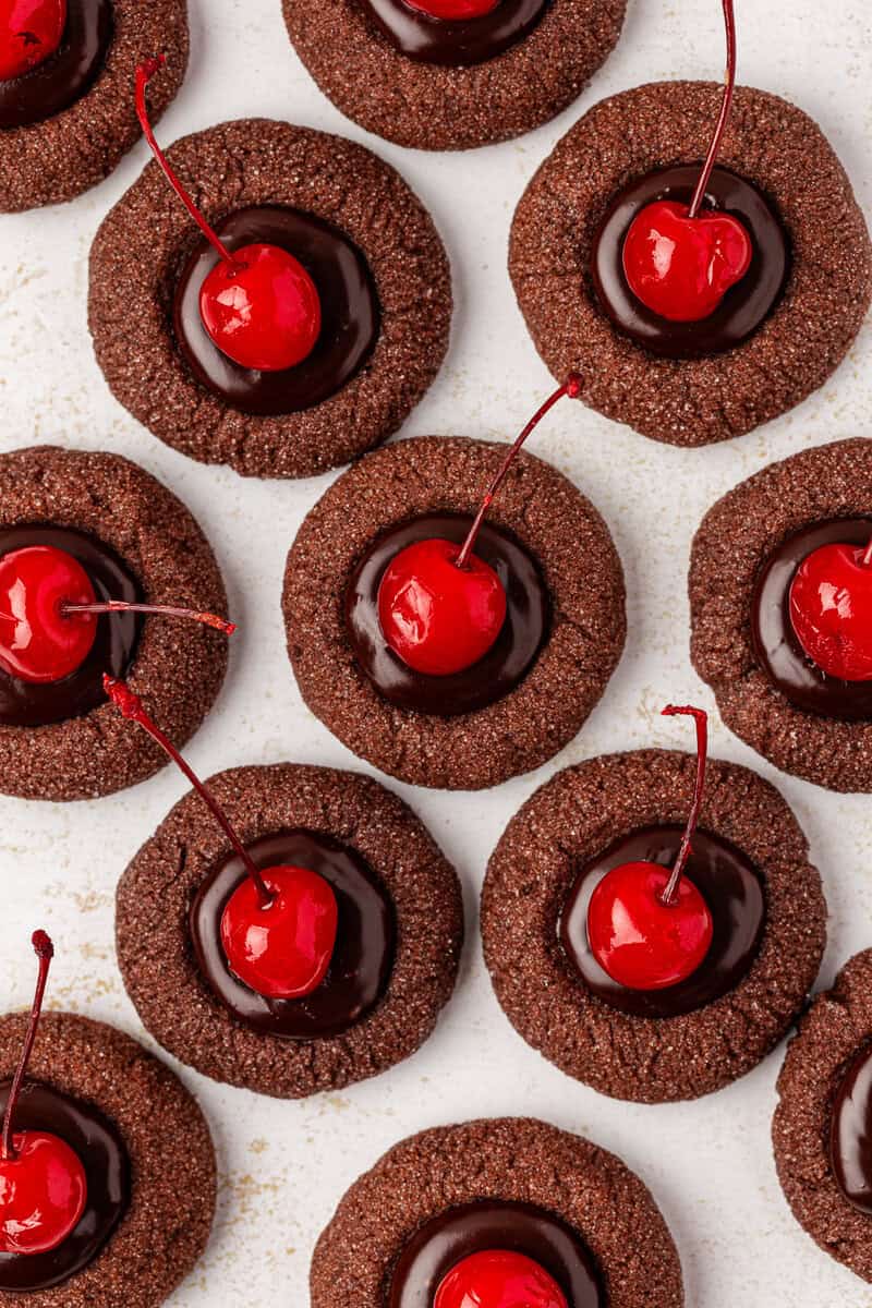 Chocolate thumbprint cookies topped with glossy chocolate filling and bright red maraschino cherries, arranged closely together on a white surface.