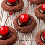Several chocolate thumbprint cookies topped with glossy chocolate ganache and bright red maraschino cherries are arranged on a cooling rack.