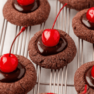 Several chocolate thumbprint cookies topped with glossy chocolate ganache and bright red maraschino cherries are arranged on a cooling rack.