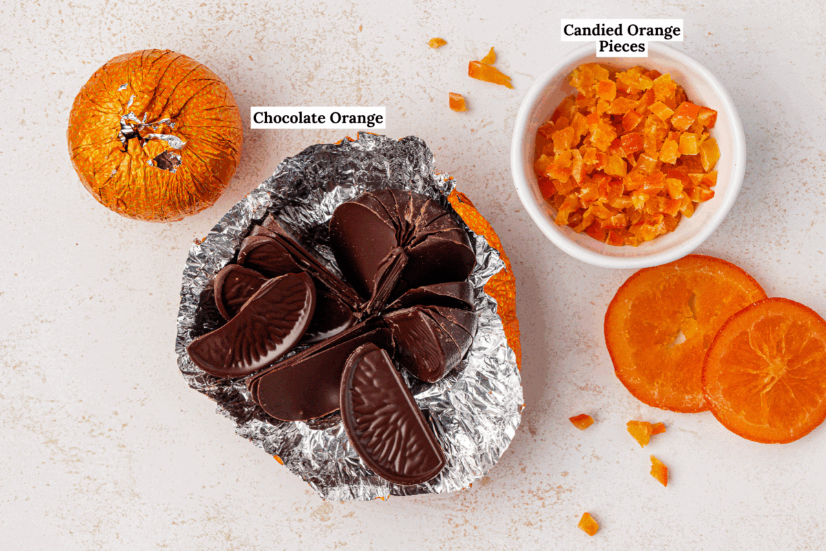 A foil-wrapped chocolate orange is partially unwrapped, showing chocolate orange slices. Nearby are candied orange pieces in a bowl, an unwrapped chocolate orange, and two slices of dried orange.