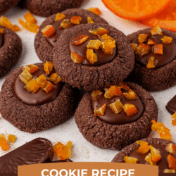 Chocolate thumbprint cookies filled with chocolate and topped with candied orange pieces are arranged on a white surface, with chocolate orange slices and fresh orange slices scattered around. Text overlay reads Chocolate Orange Thumbprint Cookies.