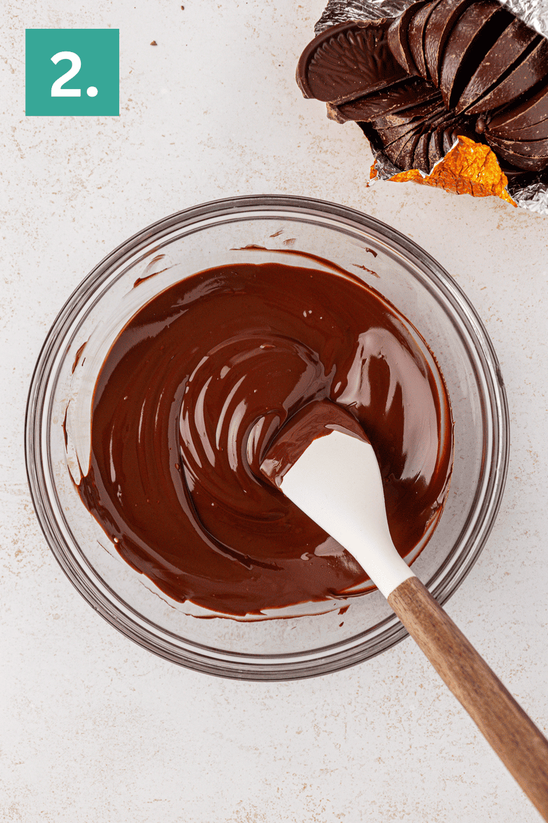 A glass bowl of melted chocolate being stirred with a white spatula. An opened chocolate orange sits in the top right corner. A green square with the number 2 is in the top left corner.