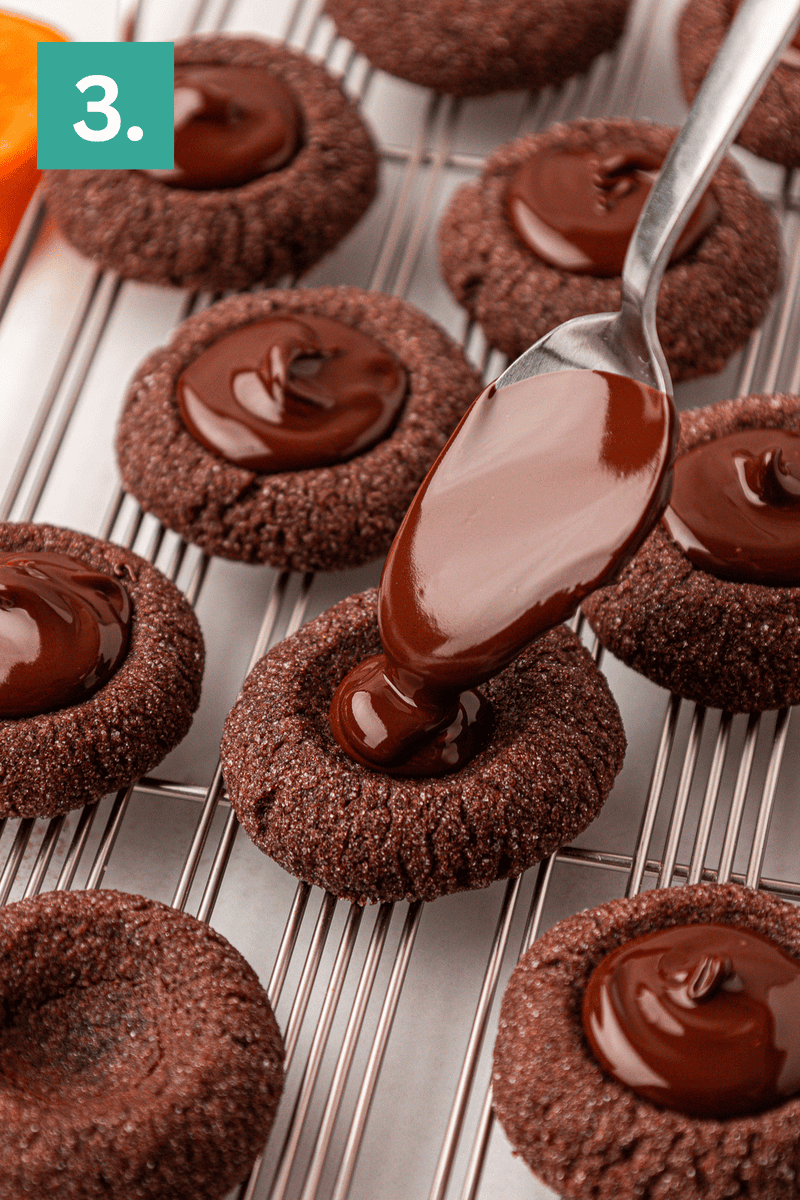A spoon drizzles glossy melted chocolate into the center of chocolate thumbprint cookies on a cooling rack. A green square with the number “3.” appears in the top left corner.