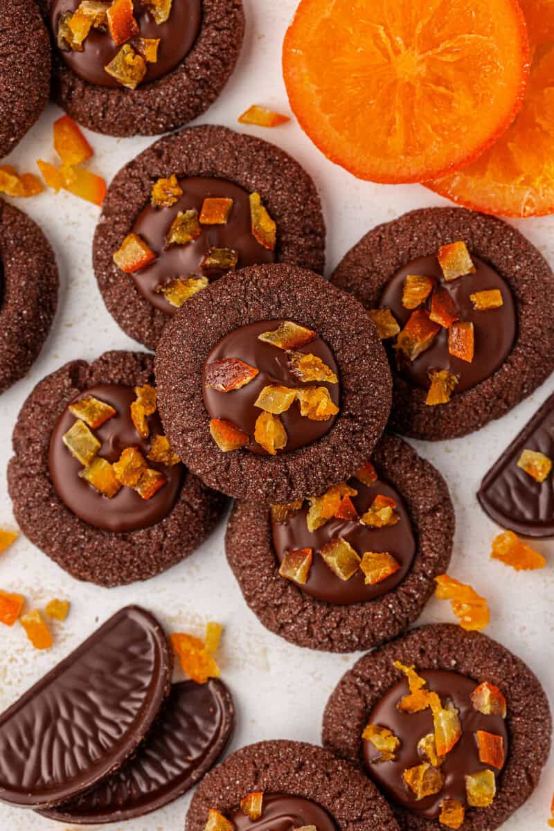 A pile of chocolate thumbprint cookies topped with chocolate and small pieces of candied orange, surrounded by chocolate orange slices and fresh orange slices on a light surface.