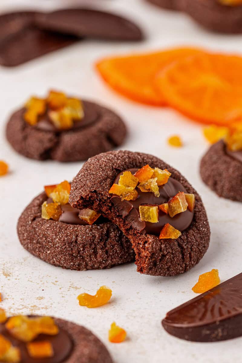 Chocolate cookies topped with chopped dried orange pieces are arranged on a light surface. One cookie is broken in half, revealing its soft inside. Slices of dried orange and chocolate pieces are visible in the background.