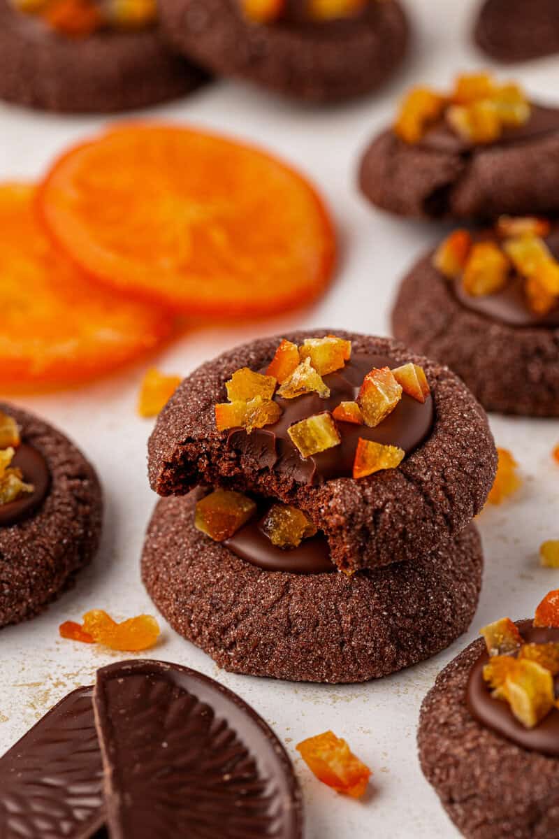 A stack of chocolate cookies topped with chocolate and candied orange pieces, with one cookie bitten. Slices of dried orange and more cookies are scattered around on a white surface.