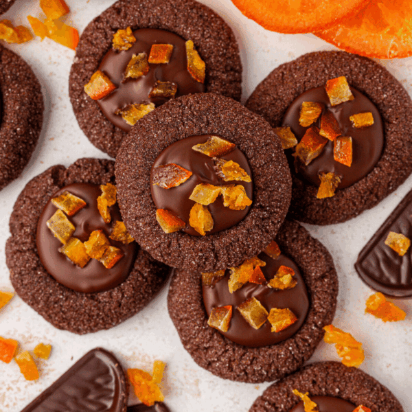 Chocolate thumbprint cookies filled with glossy chocolate and topped with small pieces of candied orange, arranged closely together with scattered orange slices and chocolate pieces nearby.