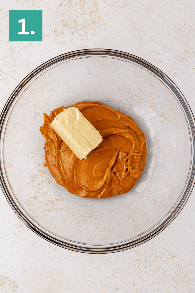 A glass mixing bowl containing creamy peanut butter and a chunk of butter. A green box with the number 1. is in the top left corner, indicating the first step of a recipe.