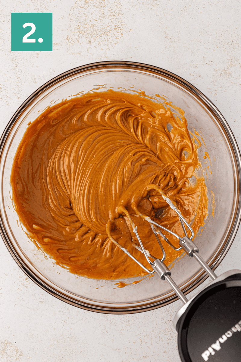 A glass bowl filled with creamy light brown batter being mixed with an electric hand mixer, shown from above. A green square with the number 2 appears in the top left corner.