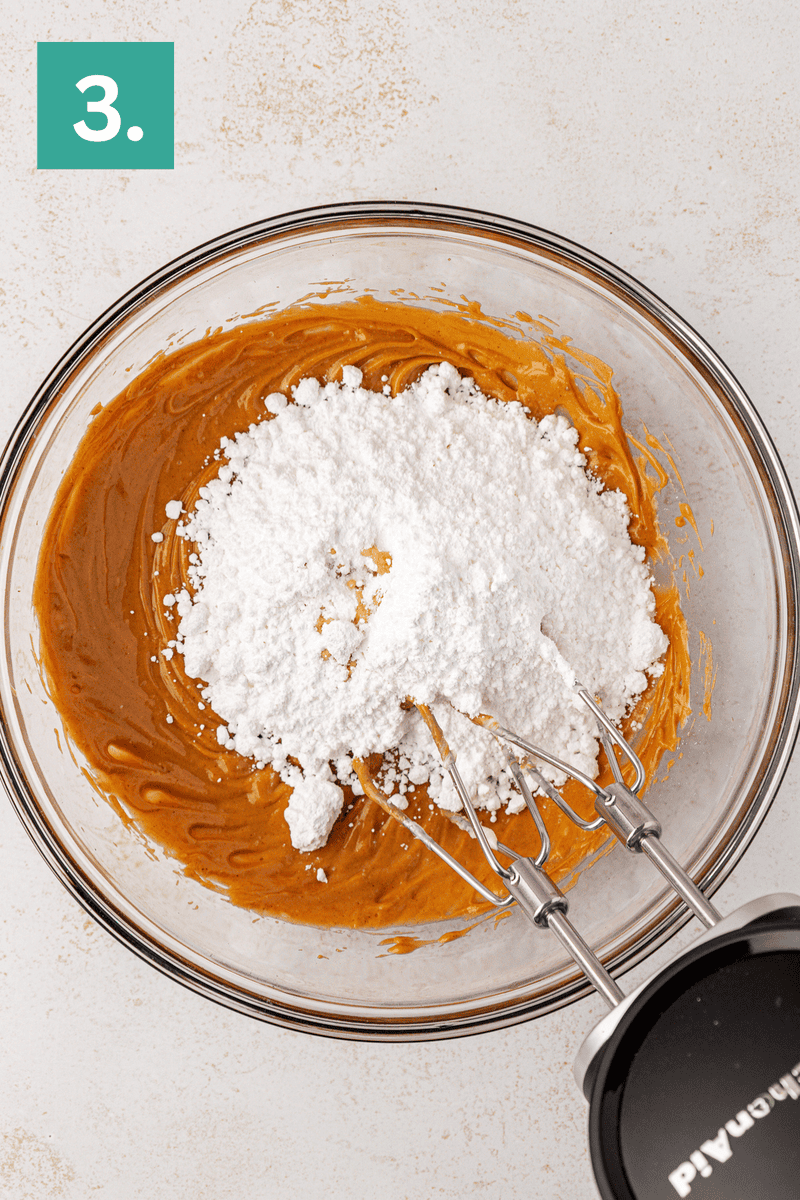 A glass bowl containing peanut butter mixture, topped with powdered sugar, is being mixed with a hand mixer. A green box with the number 3 appears in the top left corner.