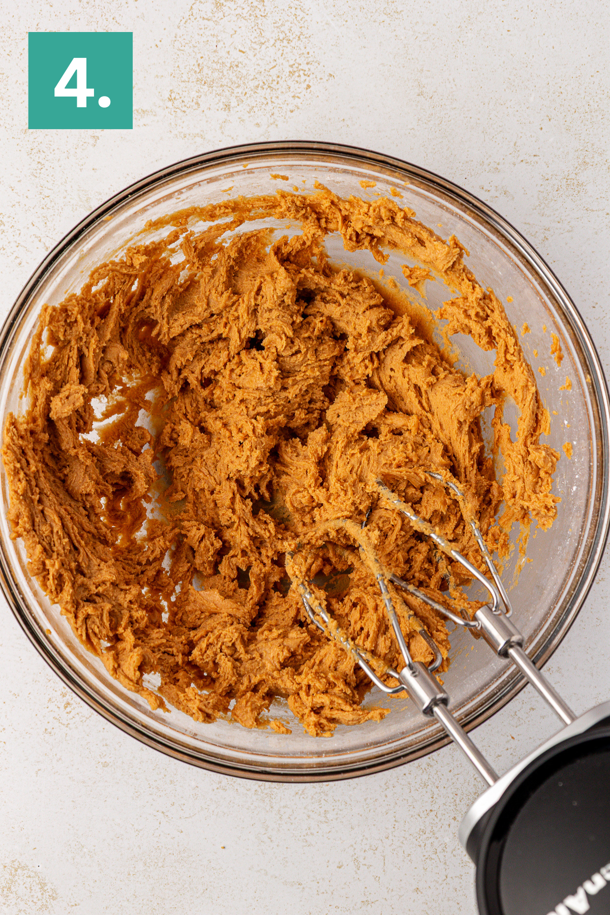 A glass bowl filled with light brown cookie dough being mixed with an electric hand mixer; a green square labeled 4. is in the top left corner.