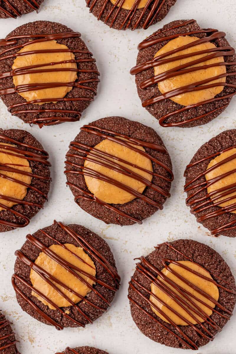 Chocolate cookies topped with a round peanut butter center and drizzled with chocolate, arranged closely together on a light-colored surface.