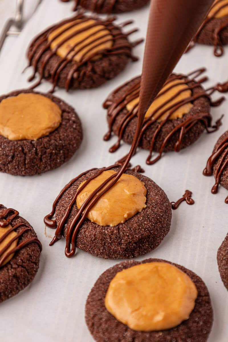 Chocolate thumbprint cookies with a peanut butter filling are arranged on parchment paper, while a piping bag is drizzling chocolate over the cookies in the foreground.