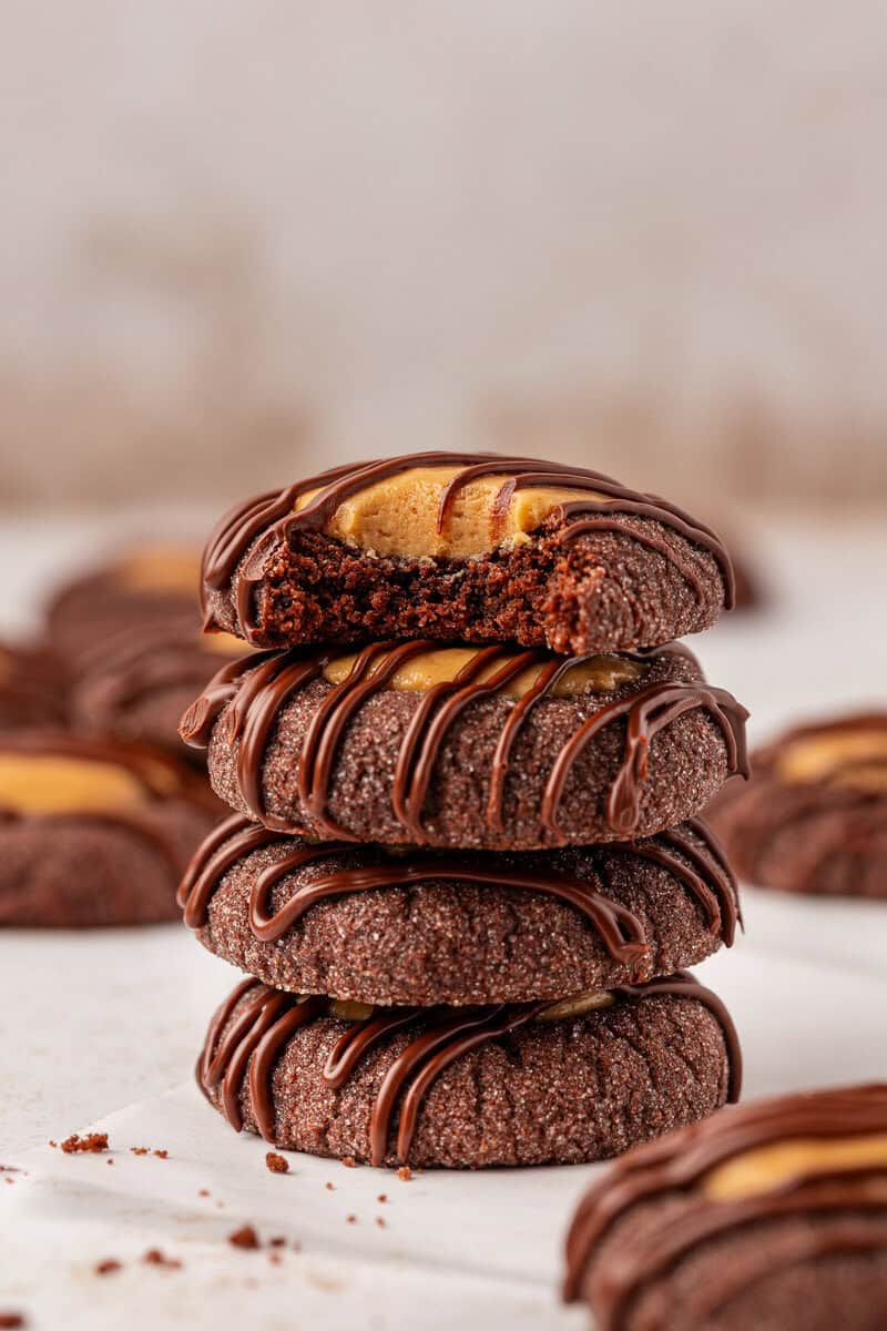 A stack of four chocolate cookies filled with peanut butter and drizzled with chocolate. The top cookie has a bite taken out, revealing the creamy peanut butter center. Crumbs are scattered on the white surface.