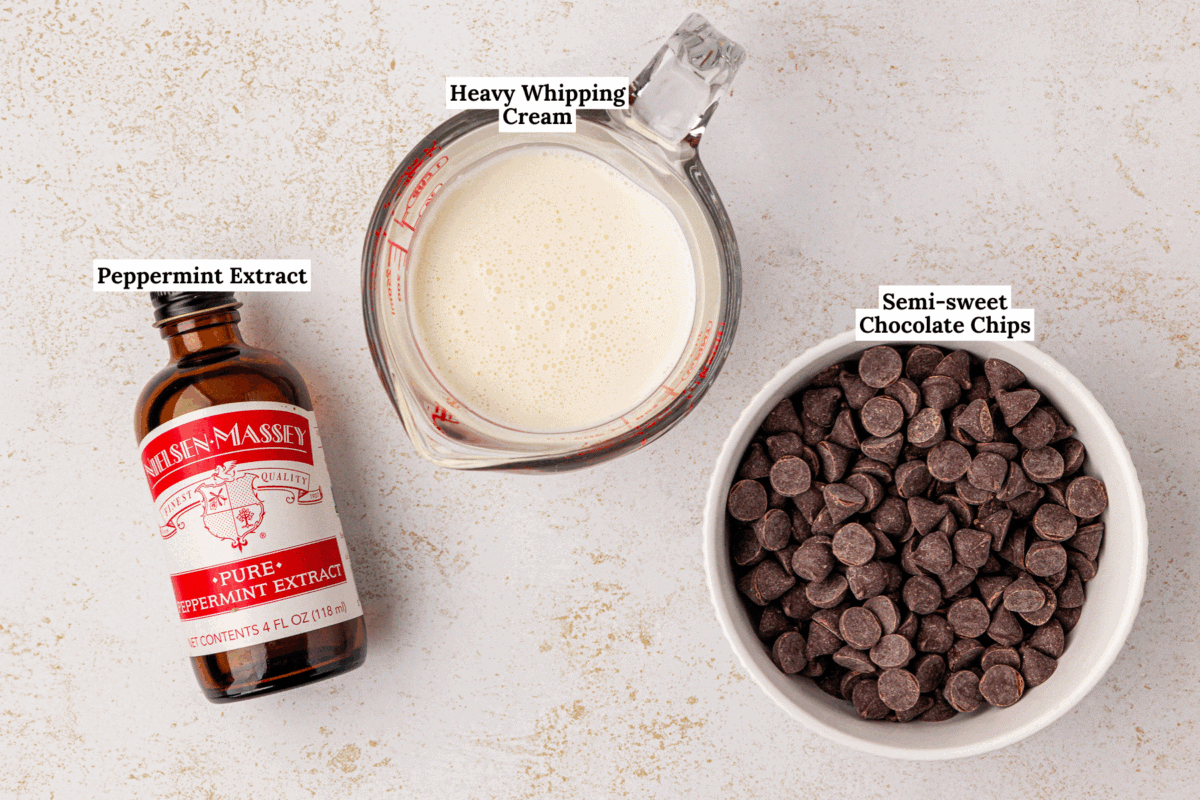 A bottle of peppermint extract, a glass measuring cup of heavy whipping cream, and a bowl of semi-sweet chocolate chips are arranged on a light countertop. Each item is labeled.
