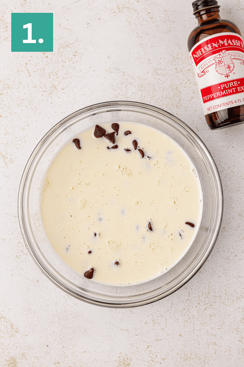 A glass bowl with milk and chocolate chips sits on a light surface. To the upper right is a bottle labeled “Peppermint Extract.” A green square with the number 1 appears in the top left corner.