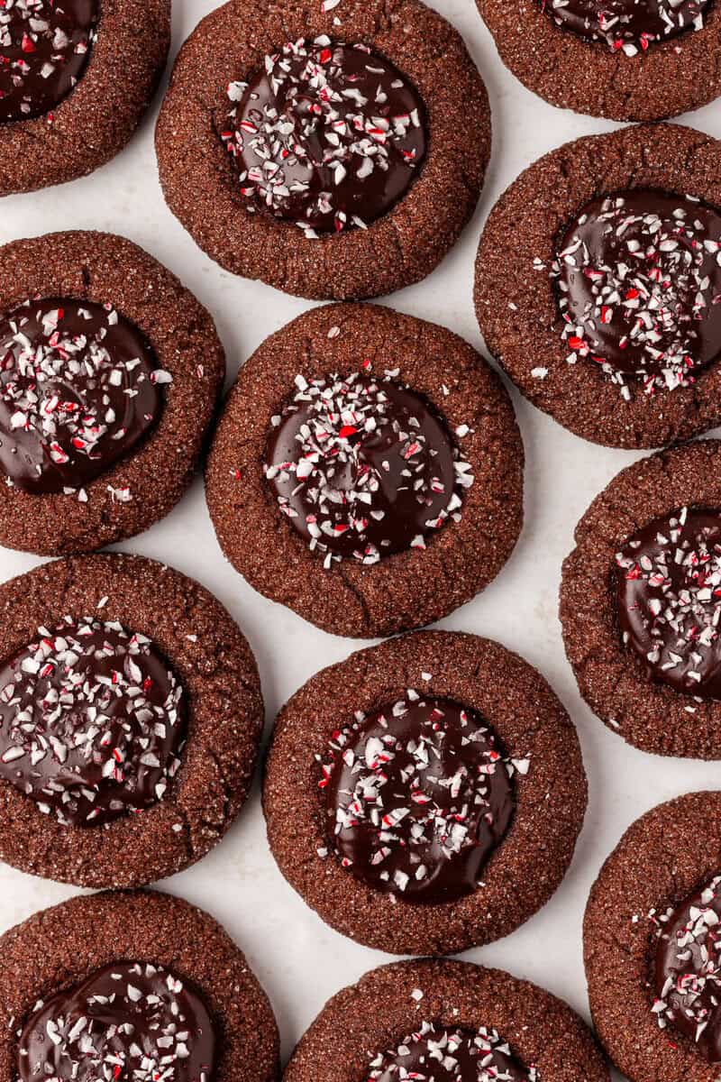 Chocolate thumbprint cookies topped with glossy chocolate centers and sprinkled with crushed peppermint candy, arranged closely together on a white surface.