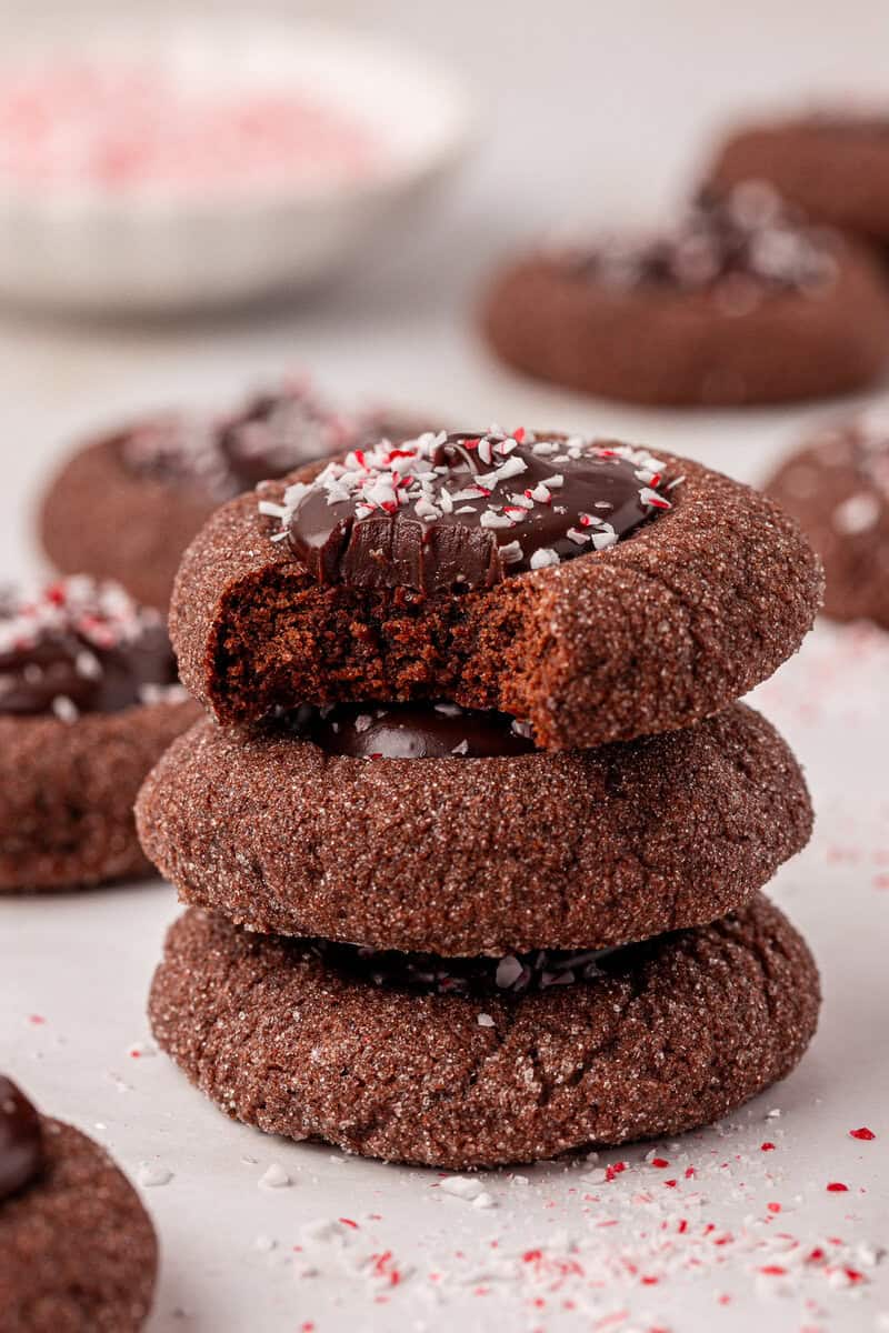 A stack of three chocolate cookies topped with chocolate and crushed peppermint, with the top cookie showing a bite taken out. More cookies and peppermint sprinkles are scattered in the background.