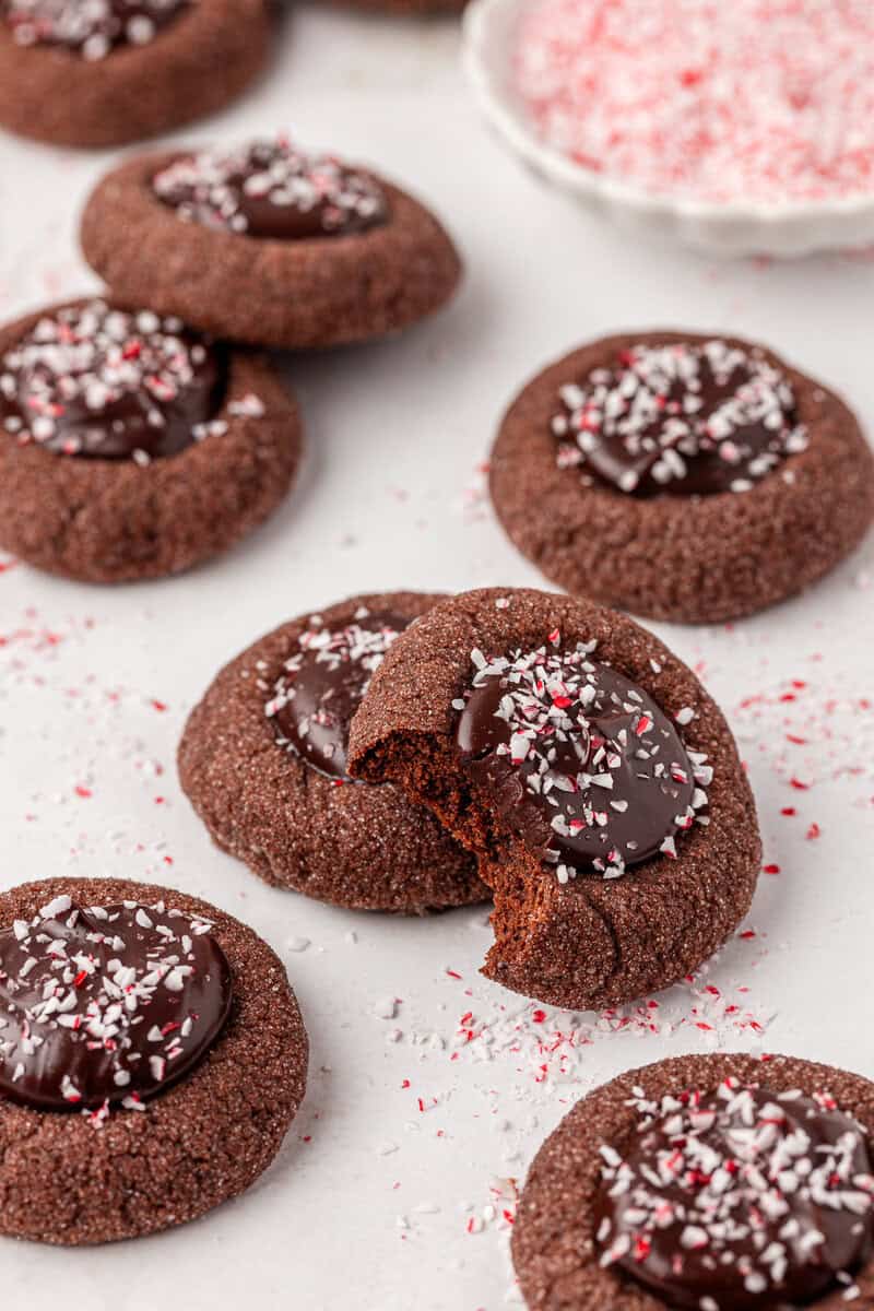 Chocolate thumbprint cookies with glossy chocolate centers are topped with crushed peppermint candy. One cookie has a bite taken out, and more cookies and peppermint pieces are scattered on a white surface.