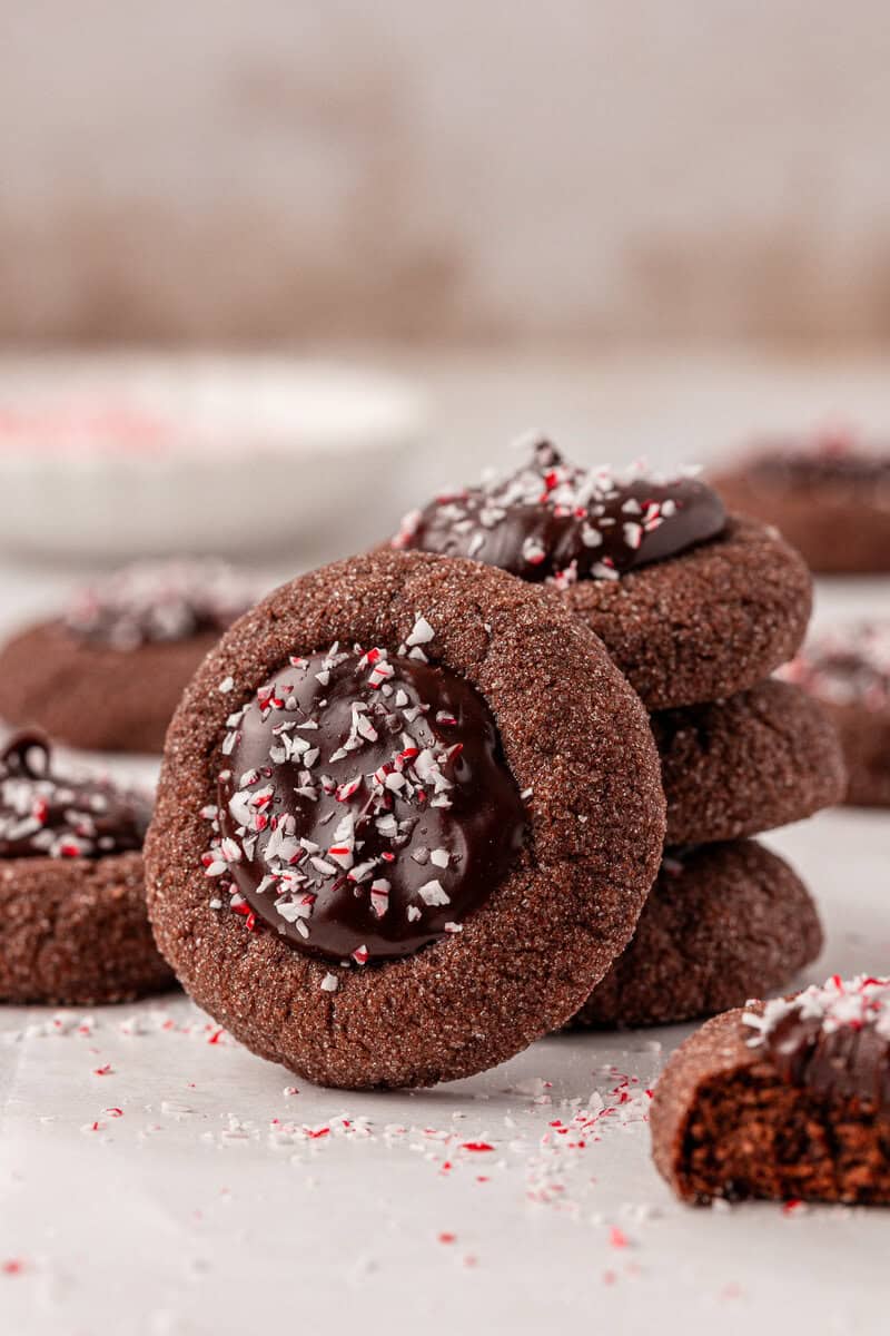 A stack of chocolate thumbprint cookies filled with melted chocolate and sprinkled with crushed peppermint, with one cookie leaning against the stack and more cookies blurred in the background.
