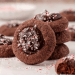 A stack of chocolate thumbprint cookies filled with glossy chocolate ganache and sprinkled with crushed peppermint pieces, with more cookies and peppermint crumbs in the background.
