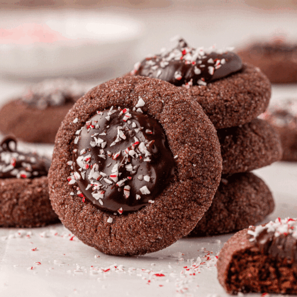 A stack of chocolate thumbprint cookies filled with glossy chocolate ganache and sprinkled with crushed peppermint pieces, with more cookies and peppermint crumbs in the background.