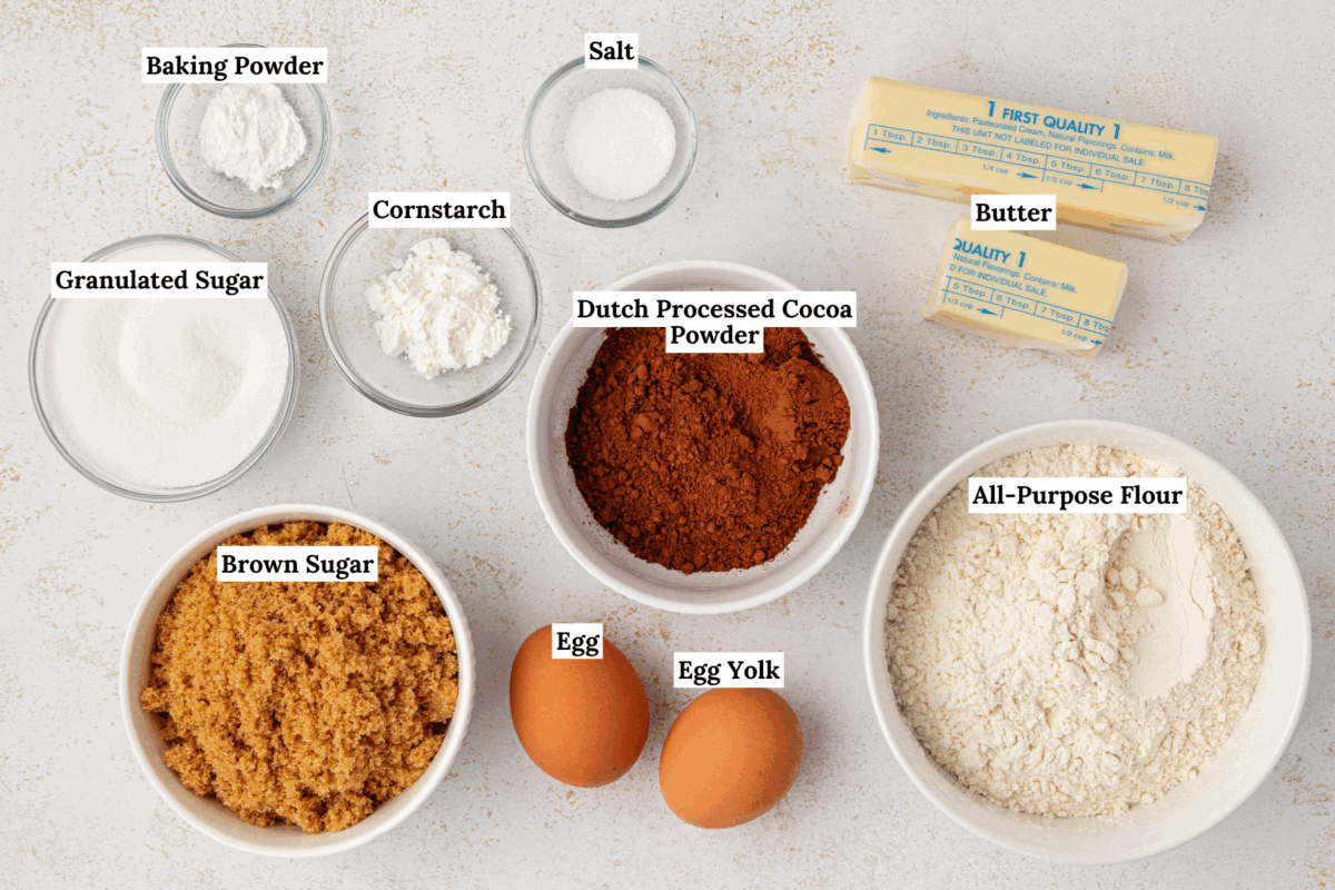 Top-down view of labeled baking ingredients in bowls on a white surface: baking powder, granulated sugar, brown sugar, cornstarch, salt, Dutch processed cocoa powder, all-purpose flour, two sticks of butter, a whole egg, and an egg yolk.