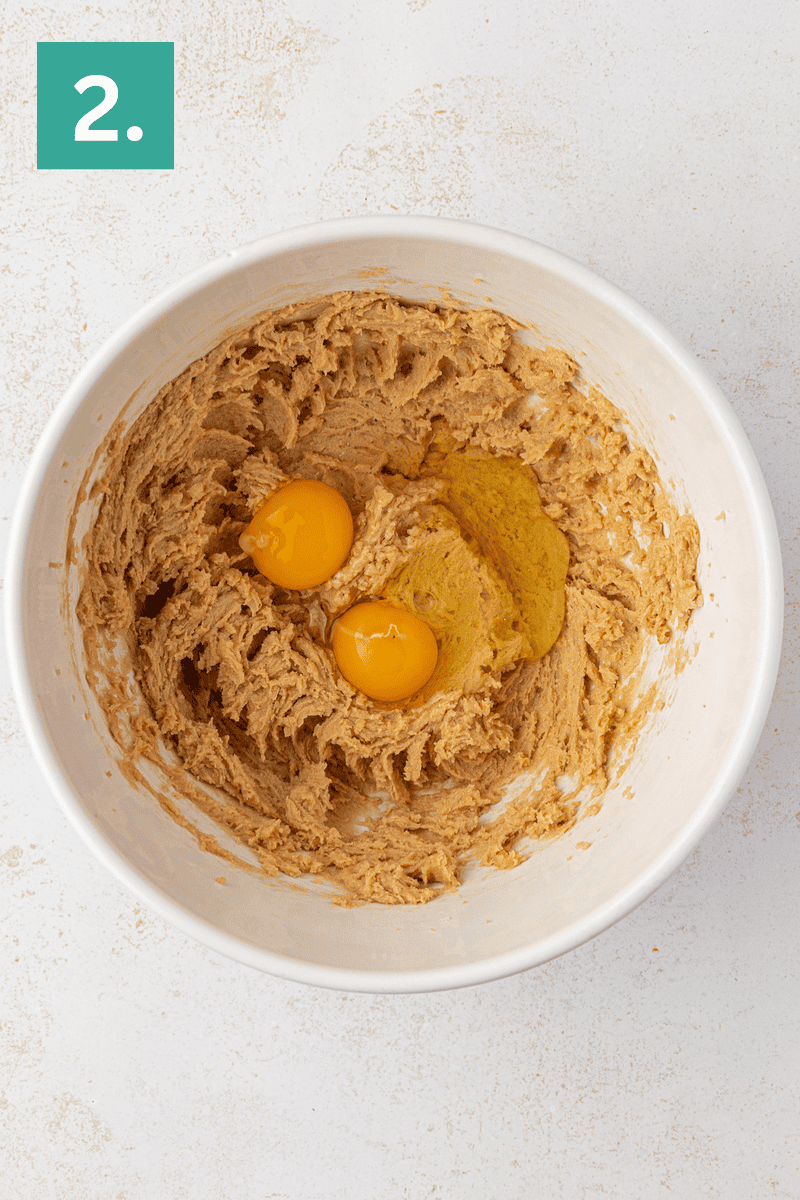 A white mixing bowl with creamed brown sugar and butter mixture, two raw eggs on top, shown from above on a light surface. A green box with the number 2 is in the top left corner.