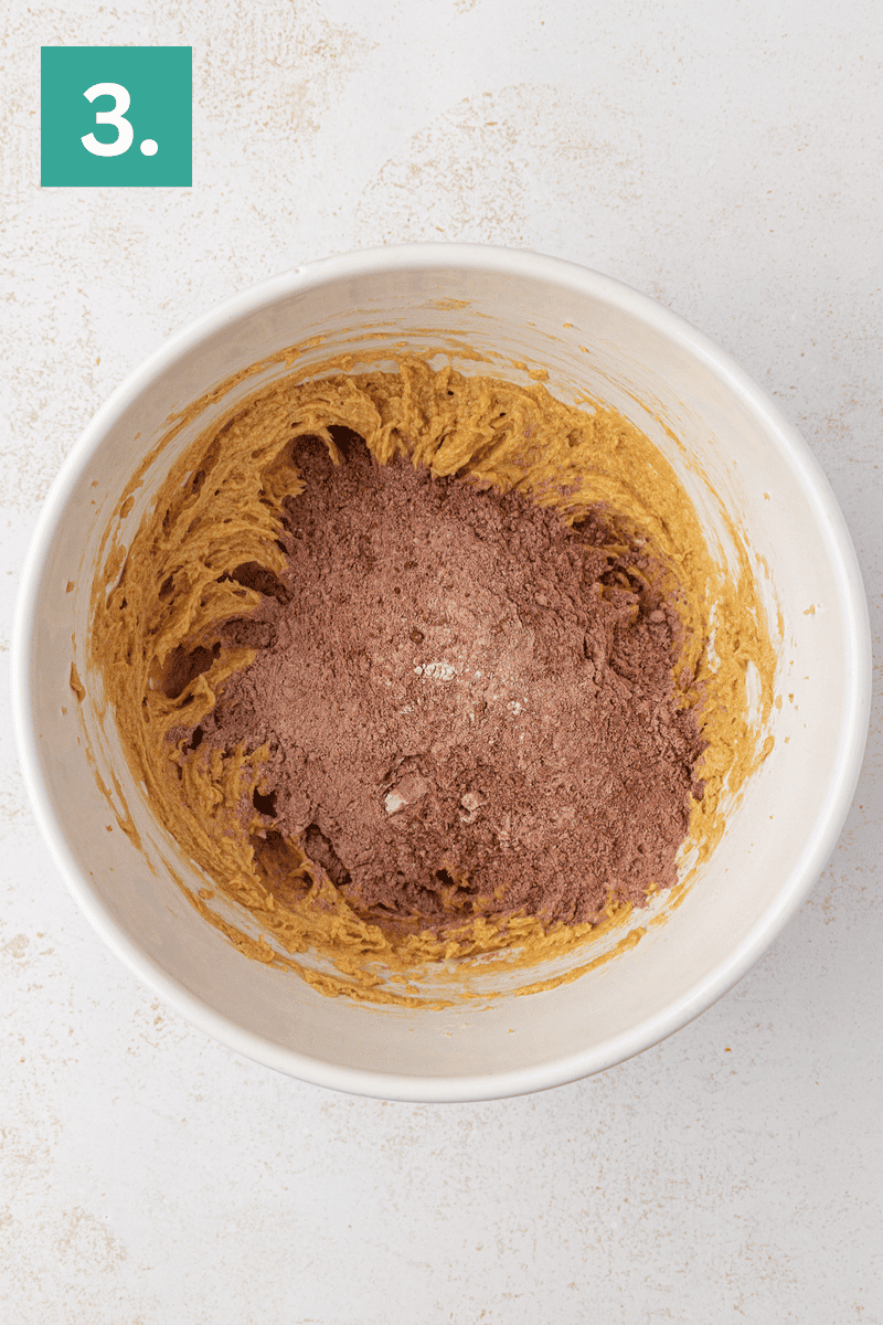 A white mixing bowl with a creamy, light brown batter partially mixed with cocoa powder, sitting on a light surface. A green box with the number 3 is in the top left corner.