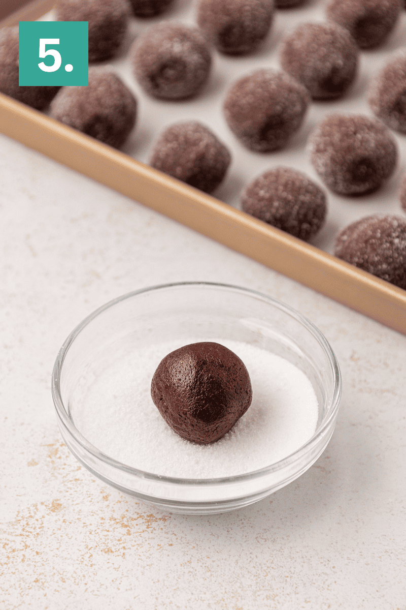 A glass bowl with a chocolate dough ball sitting in sugar, ready to be coated. In the background, a baking tray holds several sugar-coated dough balls. A green square with the number 5 is in the top left corner.