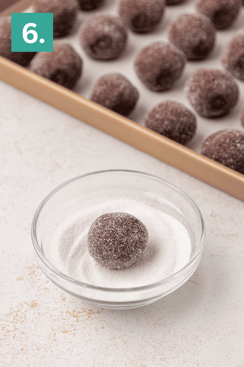 A chocolate dough ball is being coated in sugar in a glass bowl, with more sugar-coated dough balls on a baking sheet in the background. A green box with the number 6 is in the top left corner.