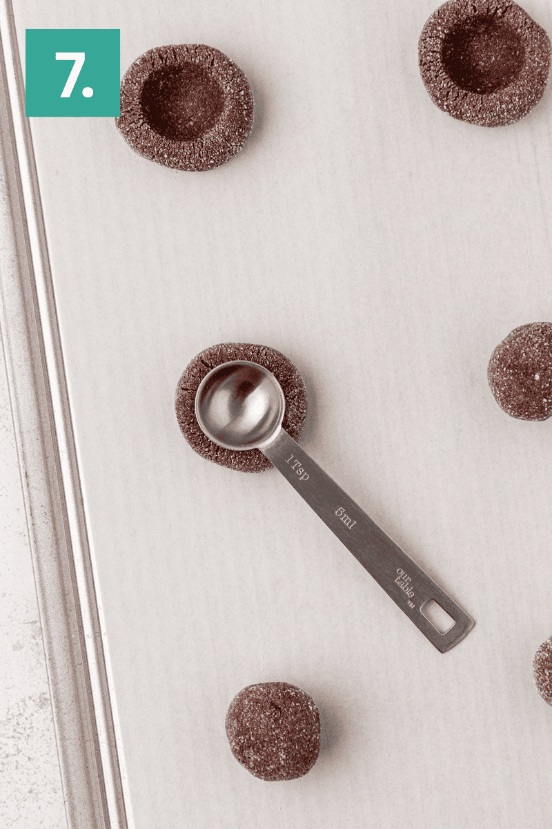 A baking tray with chocolate dough balls coated in sugar. A metal teaspoon presses an indentation into one dough ball. The number 7 is shown in a green box at the top left corner.