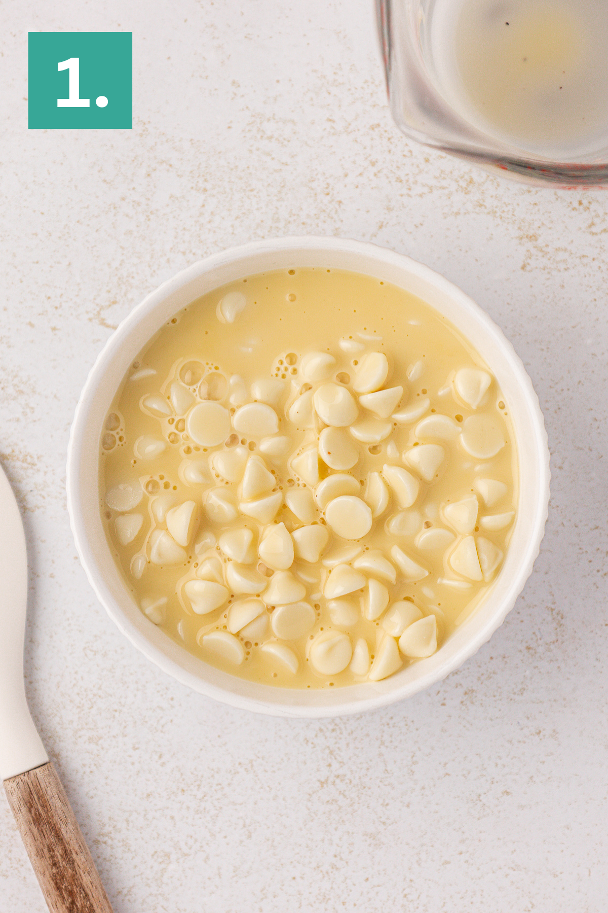 White bowl filled with yellow batter and topped with white chocolate chips, placed on a light-colored surface next to a spatula. A green box with the number “1.” is in the upper left corner.