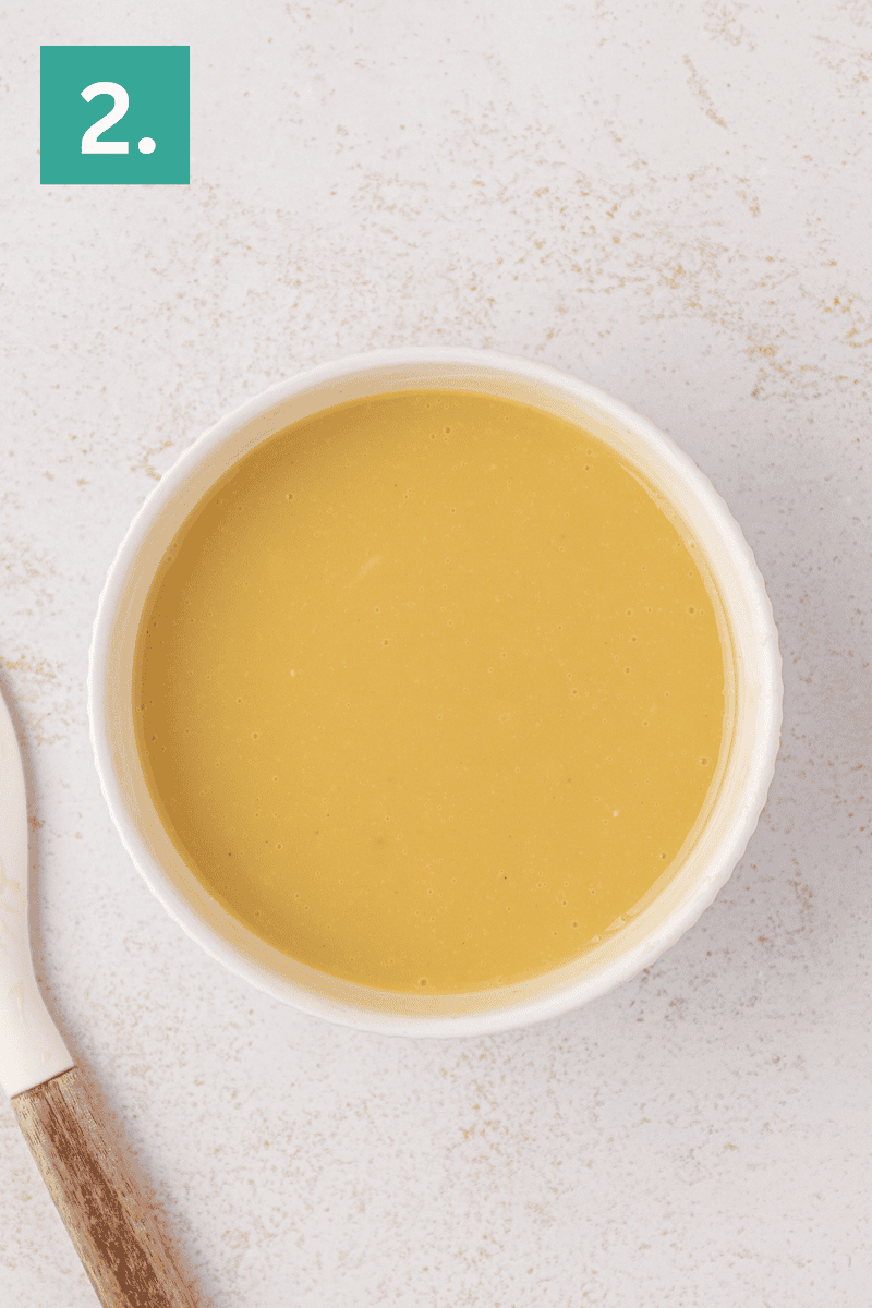 A bowl of smooth, yellow soup sits on a light-colored surface next to a utensil with a wooden handle. A green square with the number 2. is in the top left corner.
