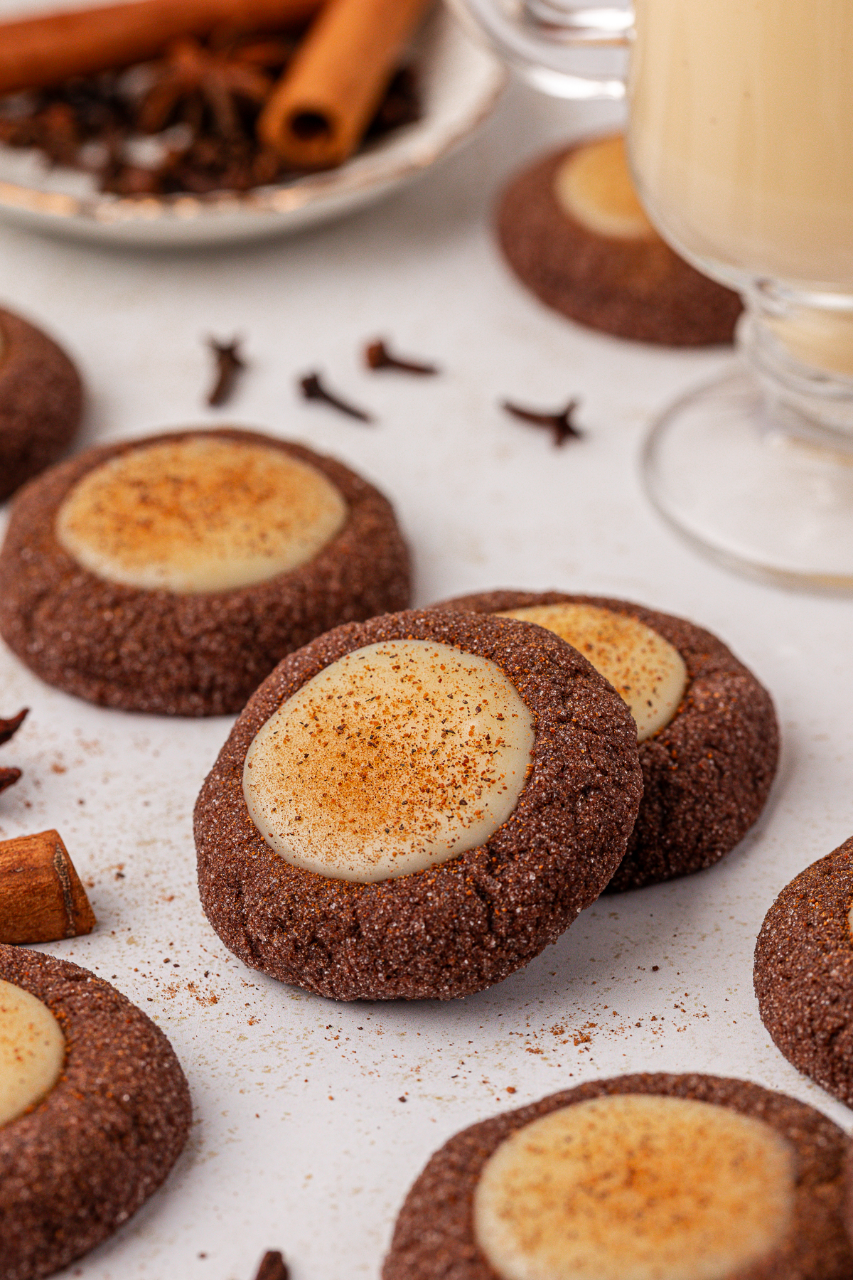 Chocolate thumbprint cookies with a creamy white center, dusted with cinnamon, are arranged on a white surface. Whole cloves and cinnamon sticks are scattered around as decoration.